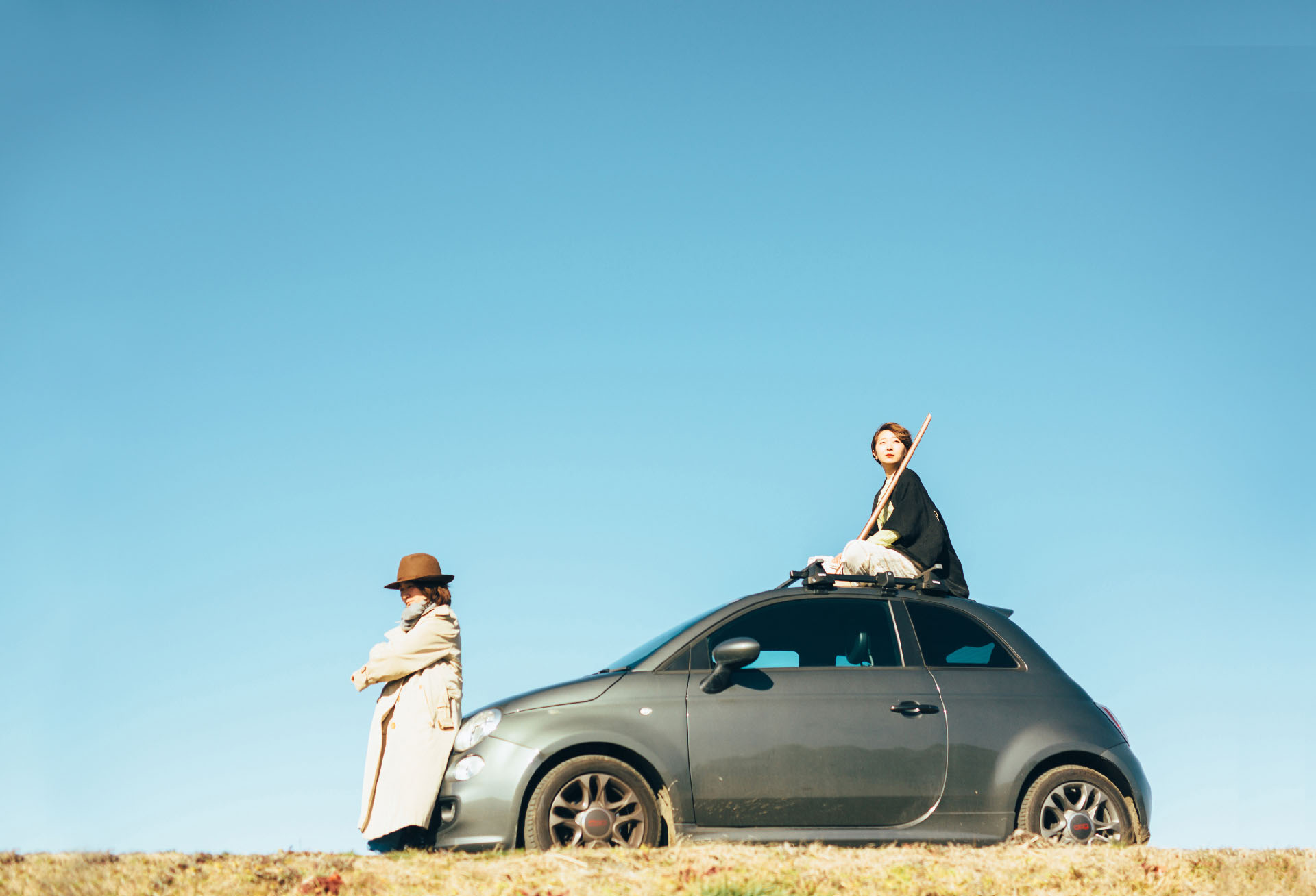 真っ青の空の下、ルパン三世を思い出すようなコンパクトミニカーの上に乗る一人の女性。その前には茶色のハットを着用し、トレンチコートを着る女性が車に腰をかけている。