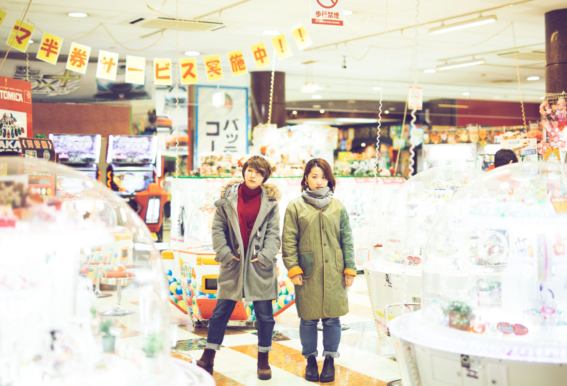 廃れたゲームセンター。それぞれのゲーム機が照明によって輝き、光の塊となっている。中心には女性2人がカメラを見つめて立っている様子。