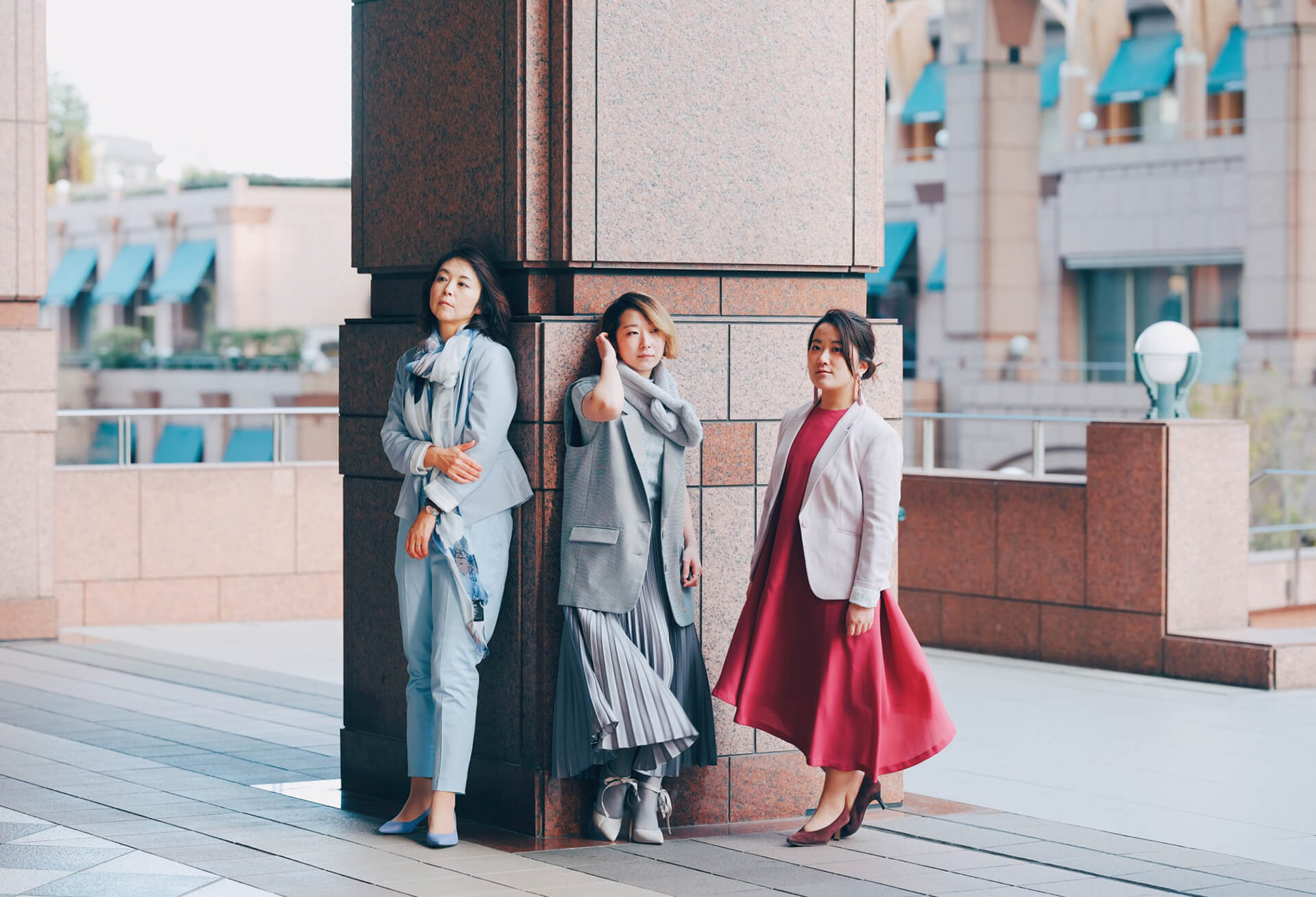 煉瓦造りの街の中、ブルーのセットアップ、グレーのワンピース、ピンクのドレスを身に纏った3人の女性がそれぞれの空を見上げて立っている様子。