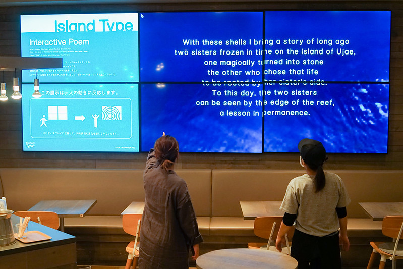 モニターに写し出されたメッセージを見る2人の女性の後ろ姿の写真