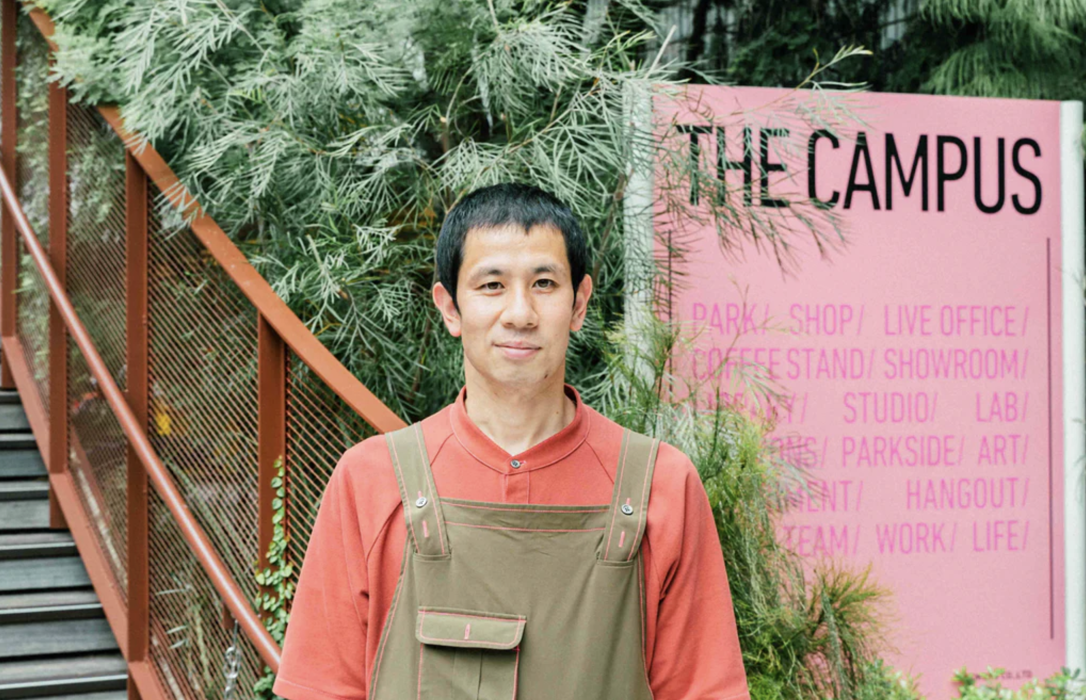 コクヨのユニフォームを着用する男性。園児色の半袖のシャツに、カーキのオーバーオール、ステッチはコクヨのカラーである傾向ピンクになっていて、自然とオフィスのデザインに馴染んでいる。