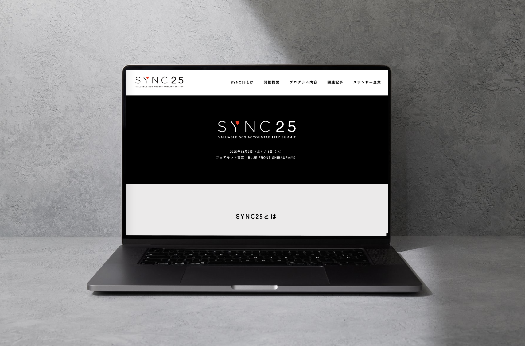 コンクリートのテーブルの上にmacが置かれ、その画面にはSYNC25の公式サイトが写っている